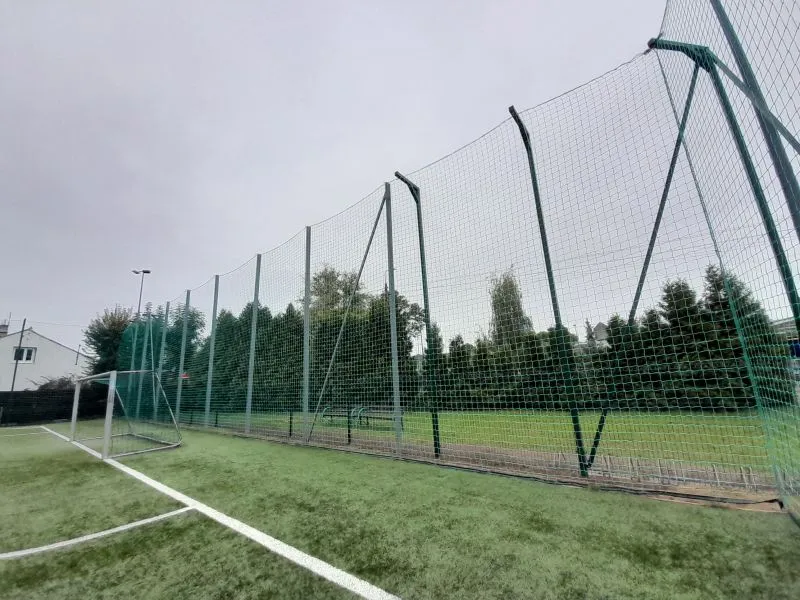 Bramka piłkarska na sztucznej nawierzchni otoczona wysoką siatką ochronną.