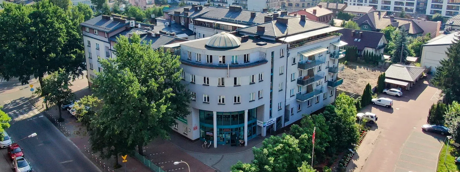 Budynek mieszkalno-usługowy z zaokrąglonym narożnikiem i szklaną fasadą na parterze otoczony drzewami, przy ruchliwej ulicy i parkingu.