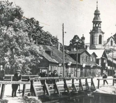 Drewniany most z kilkoma przechodniami na tle drewnianych budynków i wieży kościoła.