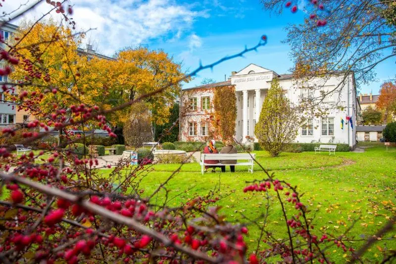 Biały budynek z kolumnami otoczony parkiem z zieloną trawą, jesiennymi drzewami i ławkami, na jednej z nich siedzą dwie osoby.