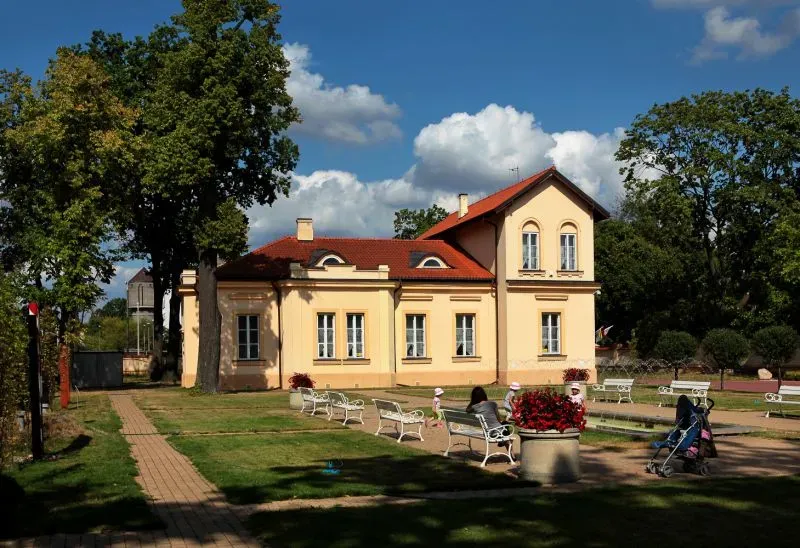 Dom z żółtą elewacją i czerwonym dachem otoczony jest trawnikiem, alejkami i ławkami, na których siedzą ludzie.