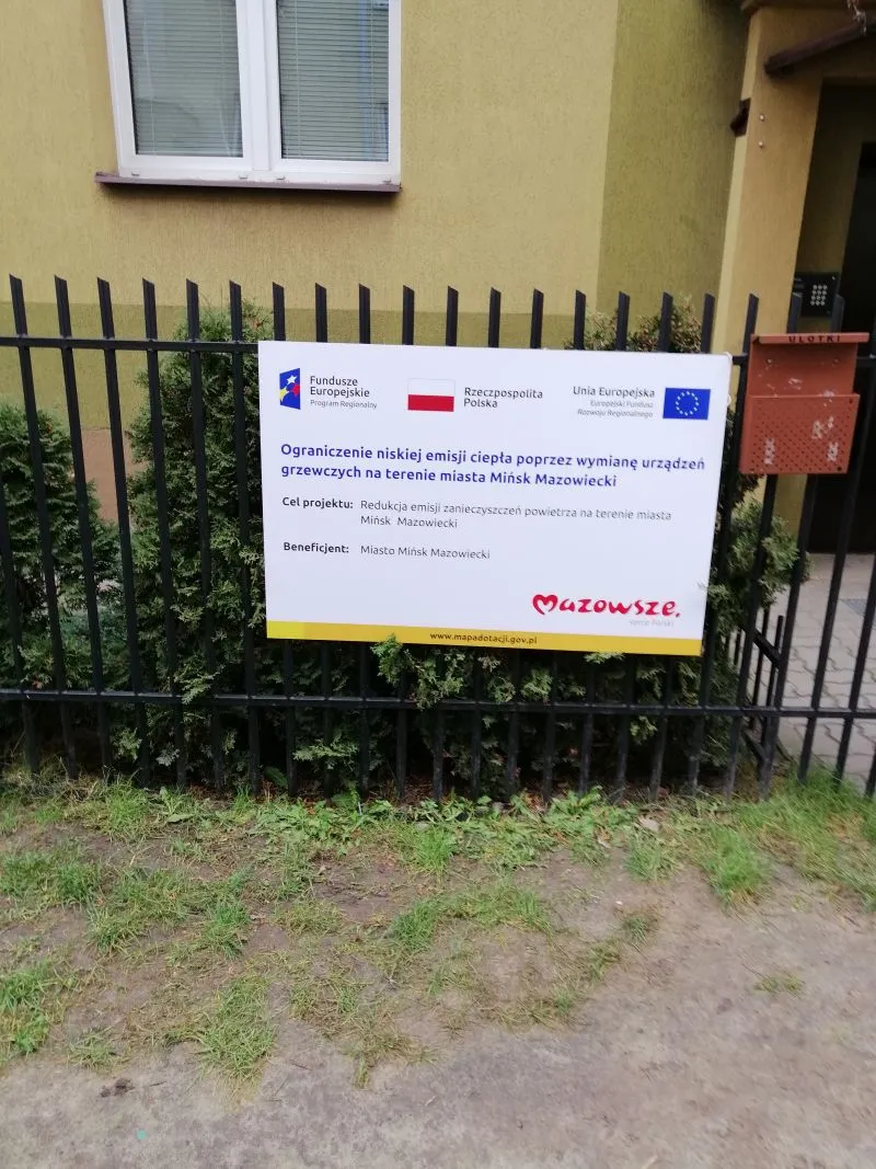 Tablica informacyjna o projekcie ograniczenia niskiej emisji ciepła poprzez wymianę urządzeń grzewczych na ogrodzeniu przed budynkiem.