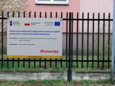 Tablica informacyjna o projekcie ograniczenia niskiej emisji ciepła poprzez wymianę urządzeń grzewczych na terenie miasta Mińsk Mazowiecki, zamocowana na czarno malowanym ogrodzeniu.