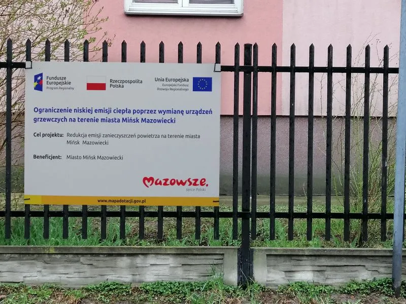 Tablica informacyjna o projekcie ograniczenia niskiej emisji ciepła poprzez wymianę urządzeń grzewczych na terenie miasta Mińsk Mazowiecki, zamocowana na czarno malowanym ogrodzeniu.