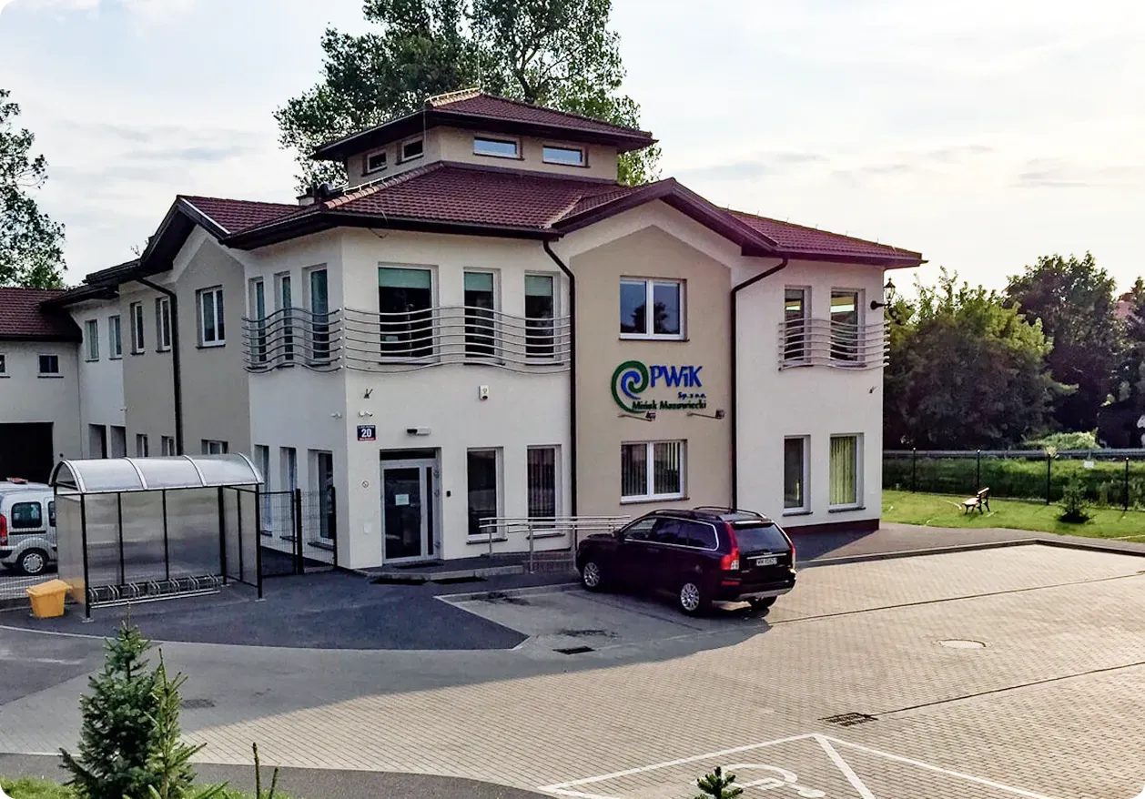Dwupiętrowy budynek z białą elewacją, czerwonym dachem, oznaczony logo PWiK oraz zaparkowany ciemny samochód na podjeździe.