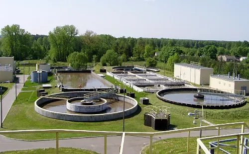 Kompleks oczyszczalni ścieków z różnymi okrągłymi i prostokątnymi zbiornikami oraz budynkami technicznymi.