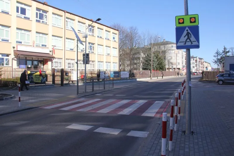 Przejście dla pieszych z sygnalizacją świetlną i znak drogowy na ulicy przy budynku szkolnym.