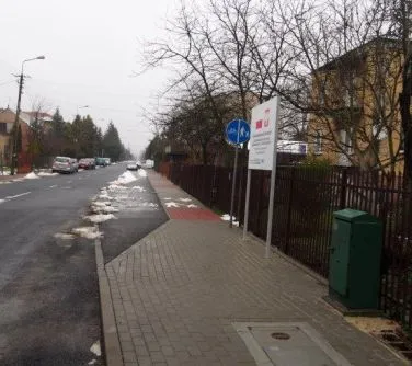 Chodnik z kostki brukowej obok ulicy z częściowo roztopionym śniegiem, przy którym stoi znak drogowy i pojemnik na śmieci.