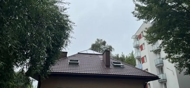 Dach domu z dwoma oknami dachowymi i kominem, otoczony drzewami, z widocznym fragmentem wielopiętrowego budynku mieszkalnego.