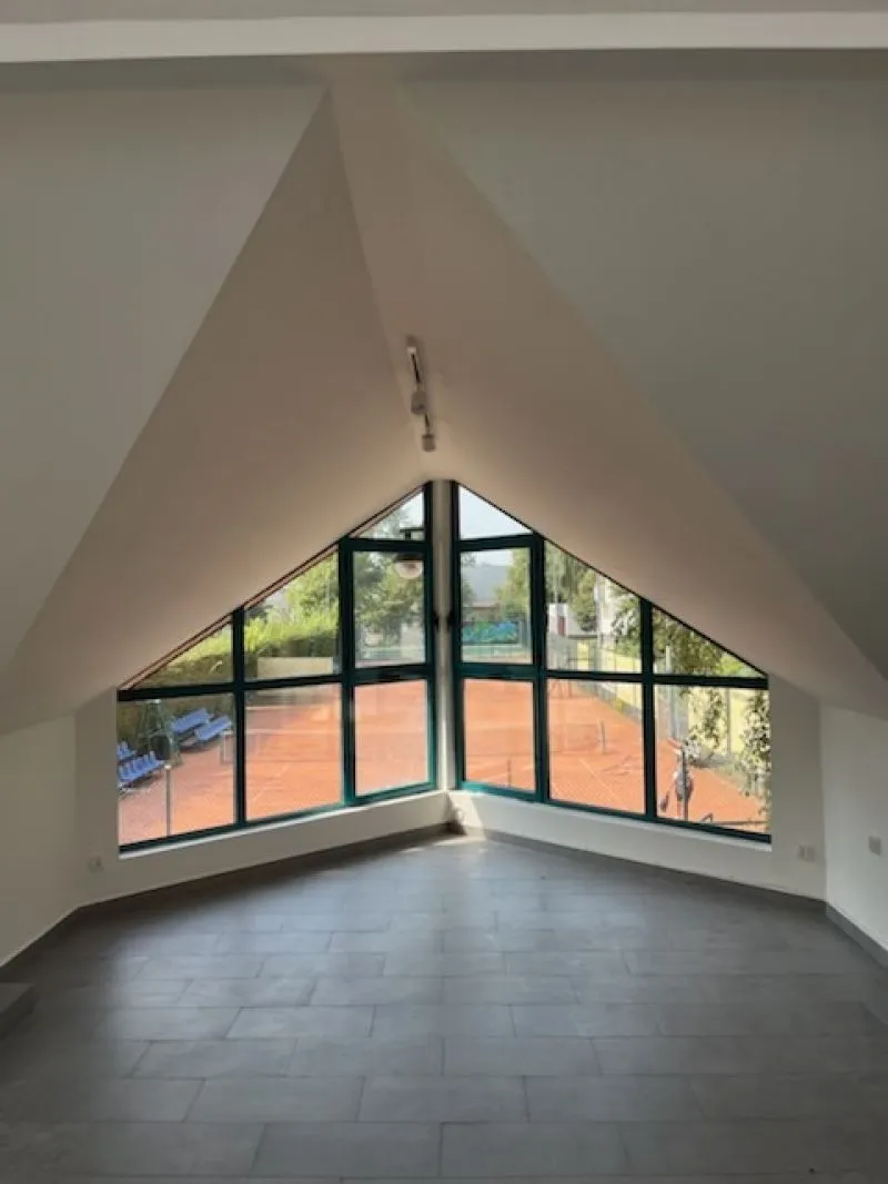 Pusty pokój z dużym trójkątnym oknem wychodzącym na kort tenisowy.