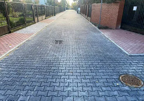 Ulica wyłożona szarymi kostkami brukowymi z dwoma włazami, po lewej stronie ogrodzenie z metalowej siatki, po prawej ceglana ściana i czarna furtka, dzień, widok wzdłuż drogi w kierunku horyzontu.