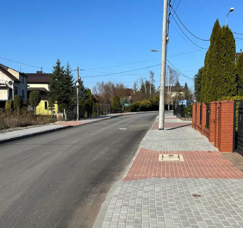 Ulica z nową nawierzchnią asfaltową i chodnikami z szaro-czerwonej kostki brukowej przy zabudowie jednorodzinnej.