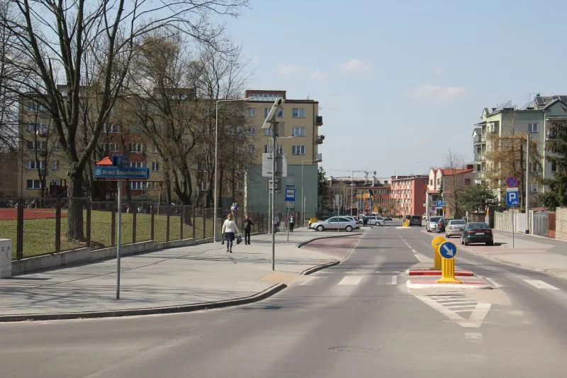 Ulica z przejściem dla pieszych, chodnikiem, kilkoma osobami, zaparkowanymi samochodami i budynkami mieszkalnymi.