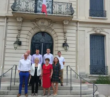 Grupa siedmiu osób stoi na schodach przed budynkiem z ozdobnymi drzwiami i balkonem z flagą.