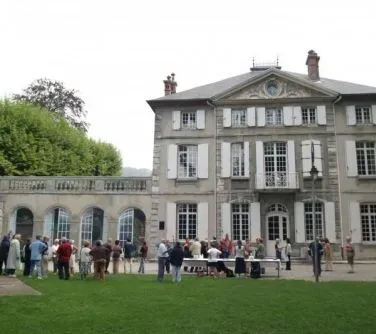 Grupa ludzi zgromadzona przed dużym, klasycystycznym budynkiem z dużymi oknami i tarasem.