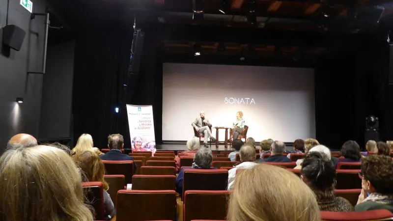 Dwóch mówców siedzi na scenie przed publicznością w sali kinowej, na ekranie za nimi wyświetlony jest napis „SONATA”.