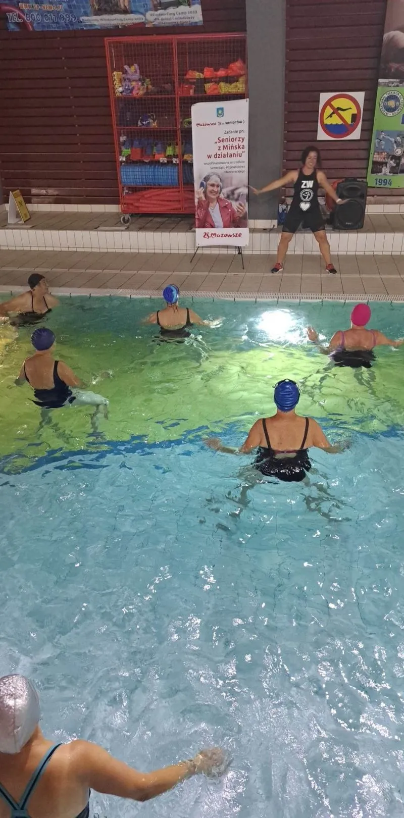 Grupa osób w czepkach kąpielowych ćwiczy w basenie pod kierunkiem instruktorki stojącej na brzegu.