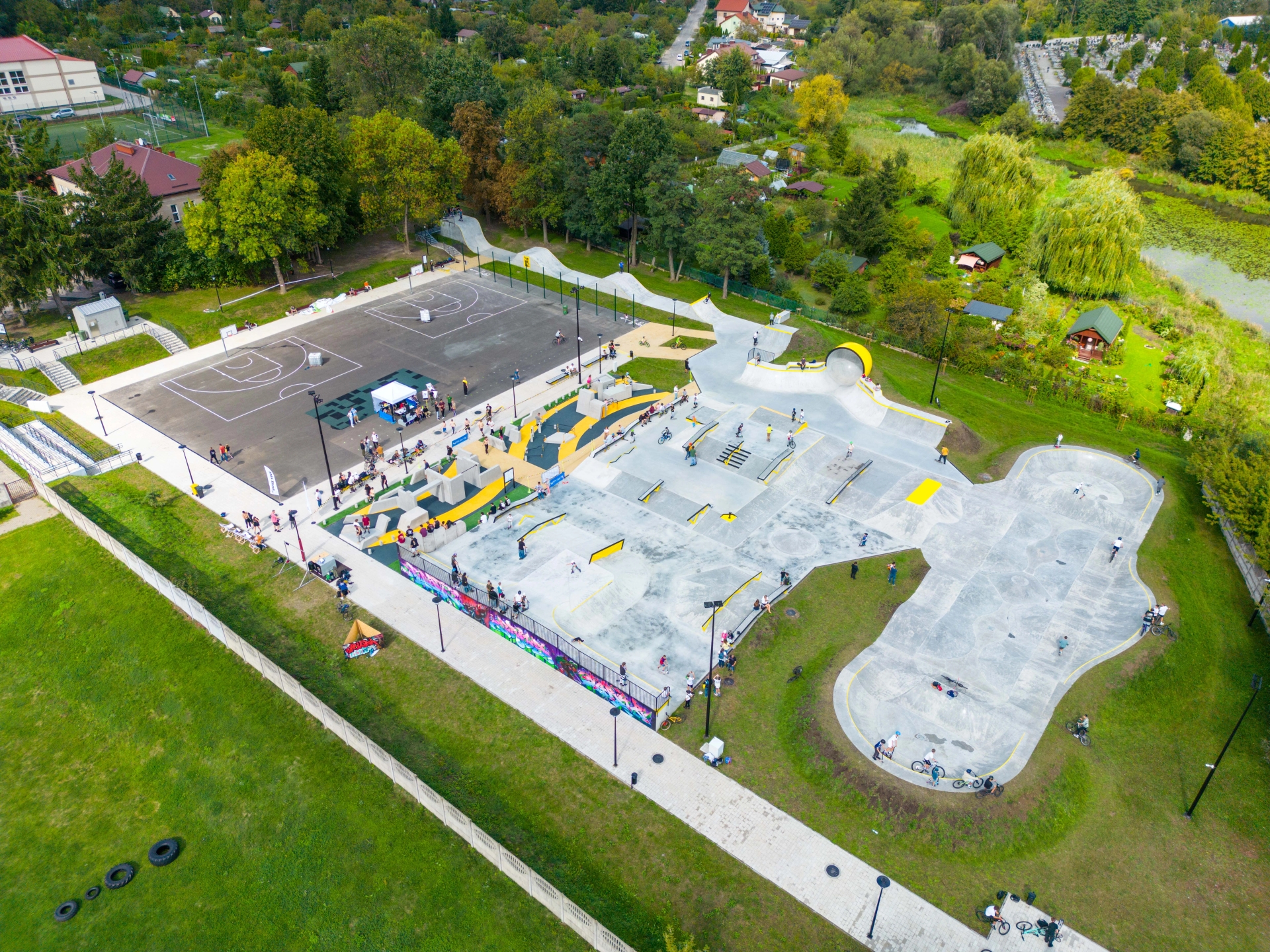 Widok z lotu ptaka na duży park sportowy w ciągu dnia. Betonowy skatepark z rampami i rowerzystami zajmuje prawą część kadru. Asfaltowe boiska do koszykówki z kilkoma osobami stoją po lewej. Otoczenie stanowią trawniki, drzewa i zabudowania w tle.