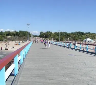 Drewniany pomost nad plażą z ludźmi spacerującymi i stojącymi przy barierkach, w tle drzewa i niebo.