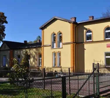 Dwupiętrowy żółty budynek z dużymi oknami i ogrodzeniem metalowym na pierwszym planie.