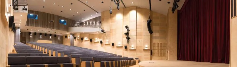 Audytorium z rzędem pustych szarych foteli i sceną oświetloną reflektorami parzącą bordową zasłonę.