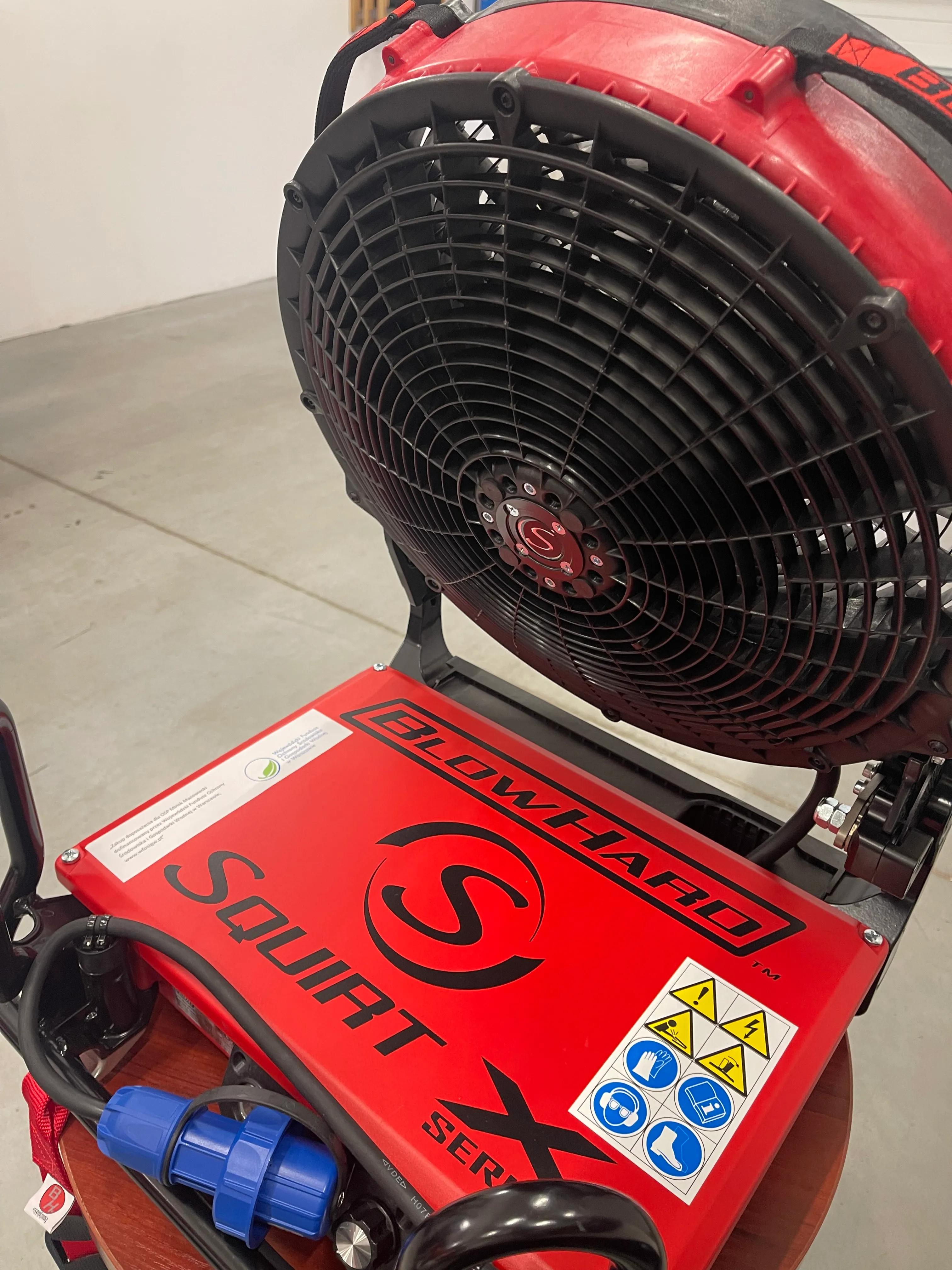 Czerwony wentylator przemysłowy z czarną ochronną kratką i logo „Squirt” na obudowie, ustawiony na szarej podłodze w pomieszczeniu.