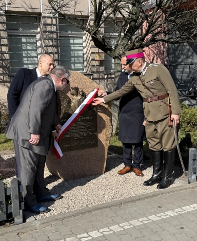 Czterech mężczyzn odsłania kamienną tablicę pamiątkową na zewnątrz w ciągu dnia. Jeden ma historyczny mundur z różowawym paskiem na czapce i szablę, pozostali noszą garnitury i płaszcze. Na tablicy widoczna metalowa płaskorzeźba orła.