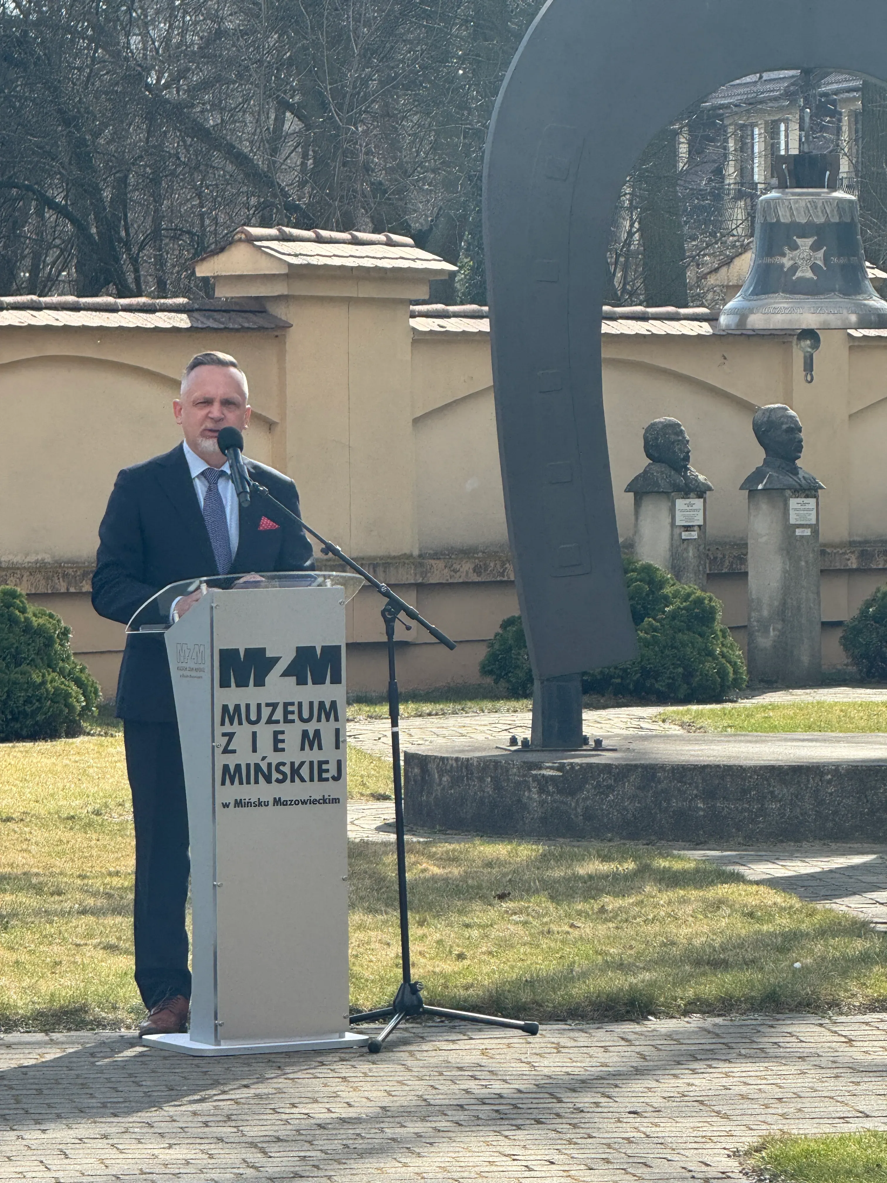 Mężczyzna w garniturze stoi za mównicą i przemawia do mikrofonu na zewnątrz w ciągu dnia. Przed nim stoi duży metalowy łuk z zawieszonym dzwonem i dwa kamienne popiersia na słupach. W tle niska beżowa ściana, drzewa i trawnik na pierwszym planie.