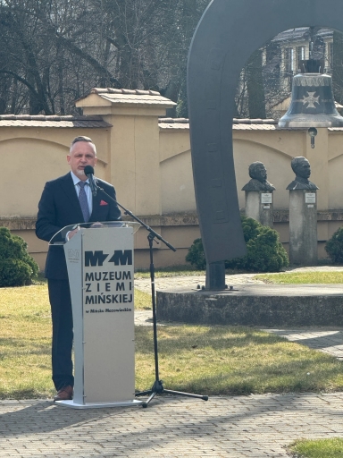 Mężczyzna w garniturze stoi za mównicą i przemawia do mikrofonu na zewnątrz w ciągu dnia. Przed nim stoi duży metalowy łuk z zawieszonym dzwonem i dwa kamienne popiersia na słupach. W tle niska beżowa ściana, drzewa i trawnik na pierwszym planie.