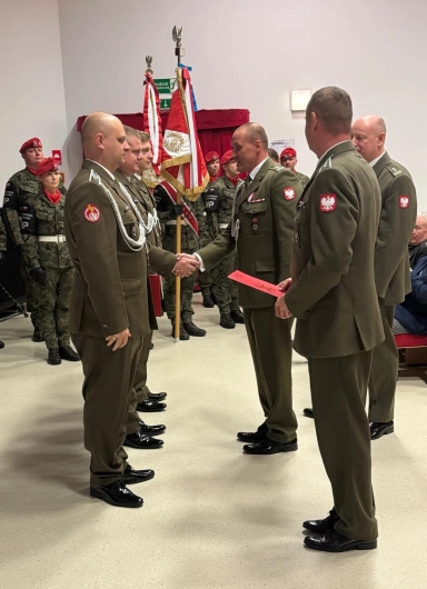 Grupa ośmiu żołnierzy w mundurach przystępuje do ceremonii wewnątrz budynku. Trzech żołnierzy stoi naprzeciwko czterech innych, dwóch z nich podaje sobie ręce. W tle znajduje się pięć osób w mundurach z czerwonymi beretami i patrolową flagą. Ściany są białe, a na podłodze jasny kolor.