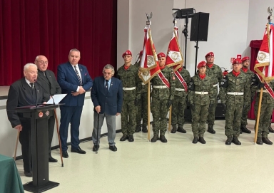 Grupa mężczyzn w garniturach i mundurach stojących w pomieszczeniu z jasną podłogą i bordową zasłoną w tle. Po lewej starszy mężczyzna przemawia przy czarnym mównicy, obok cztery osoby stojące z laskami. Po prawej osiem osób w mundurach wojskowych z czerwonymi beretami, trzy trzymają flagi.