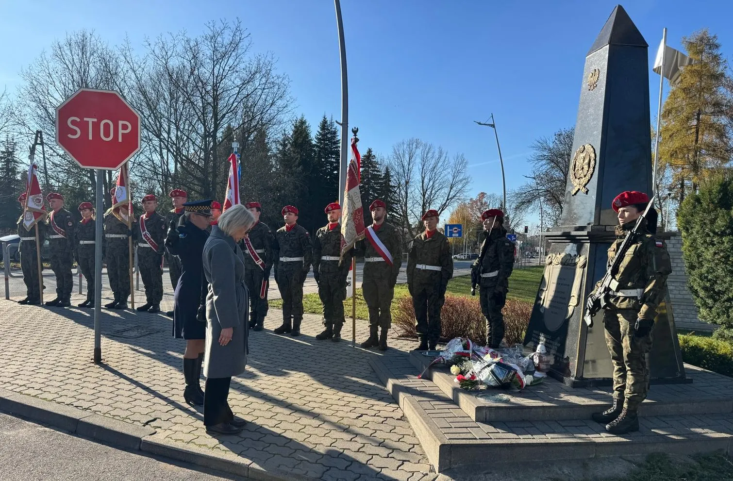 Grupa żołnierzy w mundurach z czerwonymi beretami stoi na chodniku przy pomniku. Dwie kobiety w płaszczach pochylają głowy w zadumie. Na pomniku leżą wiązanki kwiatów. Tło stanowi bezchmurne niebo i drzewa bez liści. Po lewej stronie widoczny jest znak drogowy STOP.
