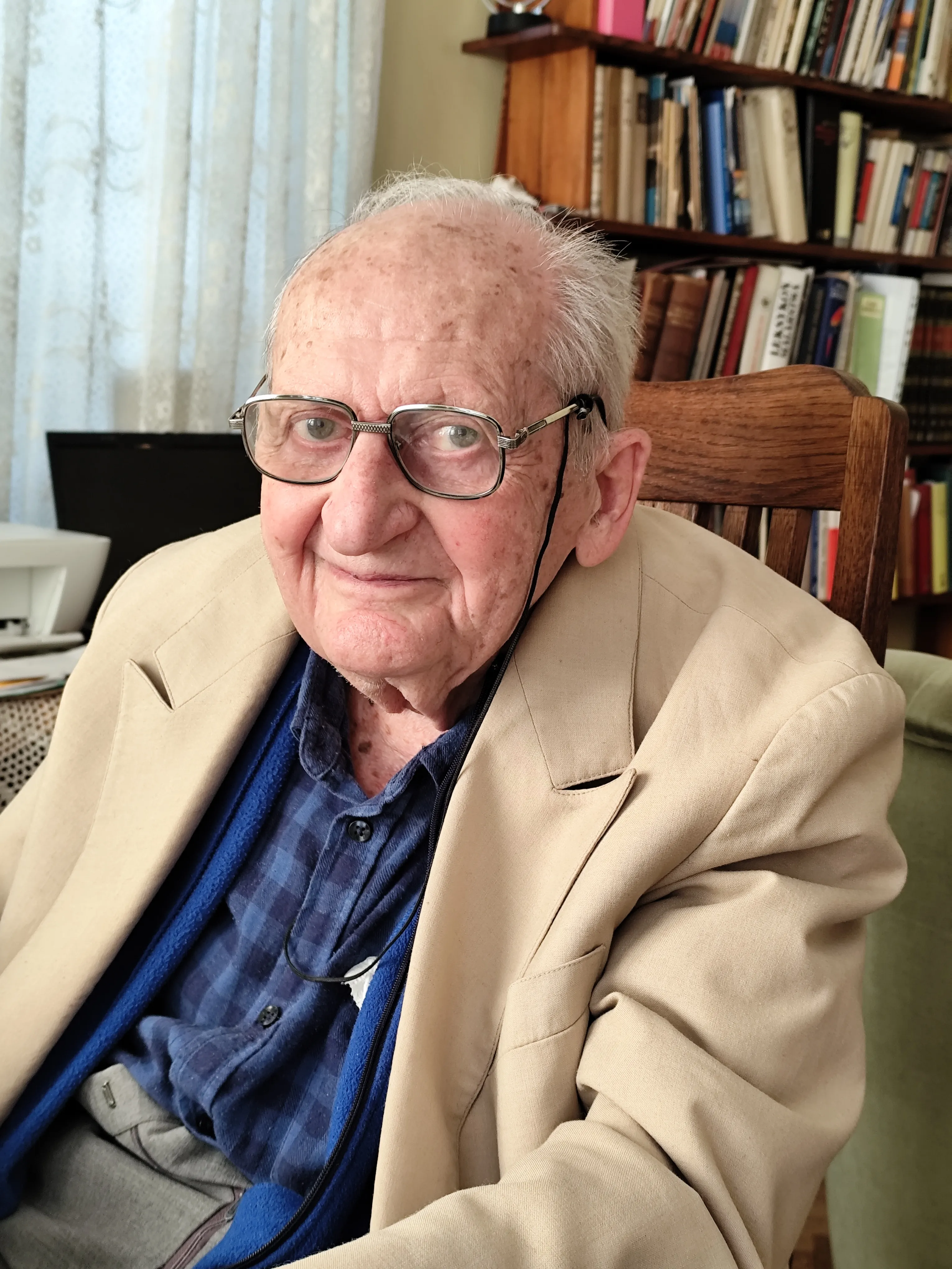 Starszy mężczyzna w beżowej marynarce i okularach siedzi na drewnianym krześle w pokoju z regałem pełnym książek.