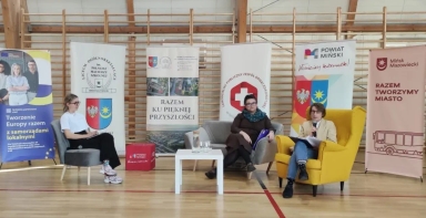 Trzy kobiety siedzą w sali gimnastycznej przed drewnianymi drabinkami. Po lewej kobieta w białej koszulce siedzi na szarym fotelu. W środku kobieta w ciemnym ubraniu siedzi na szarej sofie. Po prawej kobieta w kremowej marynarce siedzi na żółtym fotelu i trzyma mikrofon. Przed nimi stoi biały niski stolik. Za nimi stoją pionowe, kolorowe banery.