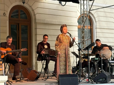 Czterech muzyków występuje na scenie na zewnątrz przy budynku z dużymi oknami, jeden śpiewa, trzech gra na instrumentach strunowych, akordeonie i perkusji.