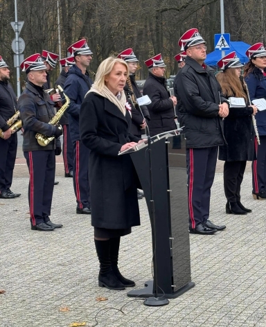 Kobieta w czarnym płaszczu i jasnym szaliku przemawia przy czarnym mównicy na zewnątrz, za nią stoi grupa muzyków w mundurach z czerwonymi czapkami, niektórzy trzymają instrumenty dęte. W tle drzewa bez liści i znak przejścia dla pieszych.