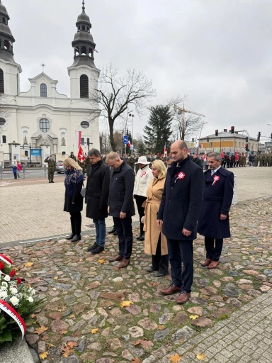 Siedem osób stoi na kamiennej nawierzchni głową skierowaną w dół, oddając hołd. W tle widać białą budowlę z dwiema wieżami oraz grupę wojskowych i flagi. Niebo zachmurzone, dzień.