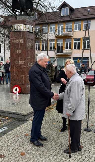 Dwóch starszych mężczyzn w kurtkach ściska sobie dłonie na zewnątrz przy pomniku z wieńcem. Obok stoi kobieta i żołnierz z bronią, w tle widoczne są budynki mieszkalne i kilka osób obserwujących scenę. Jest dzień, pora jesienna.