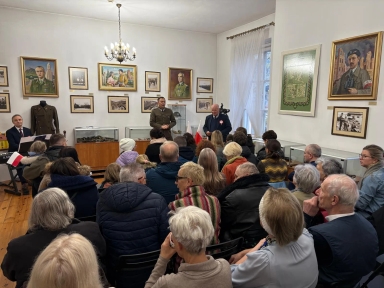 Wewnątrz jasnego pomieszczenia z białymi ścianami i drewnianą podłogą grupa ludzi siedzi na krzesłach, zwróconych w stronę dwóch mężczyzn stojących przy ścianie ozdobionej obrazami i zdjęciami historycznymi. Jeden mężczyzna ma mundur wojskowy, drugi elegancki ciemny strój z przypinką. Po lewej stronie przy pianinie siedzi kolejny mężczyzna. W pokoju wiszą obrazy z portretami i mapami, a przez okno wpada naturalne światło.