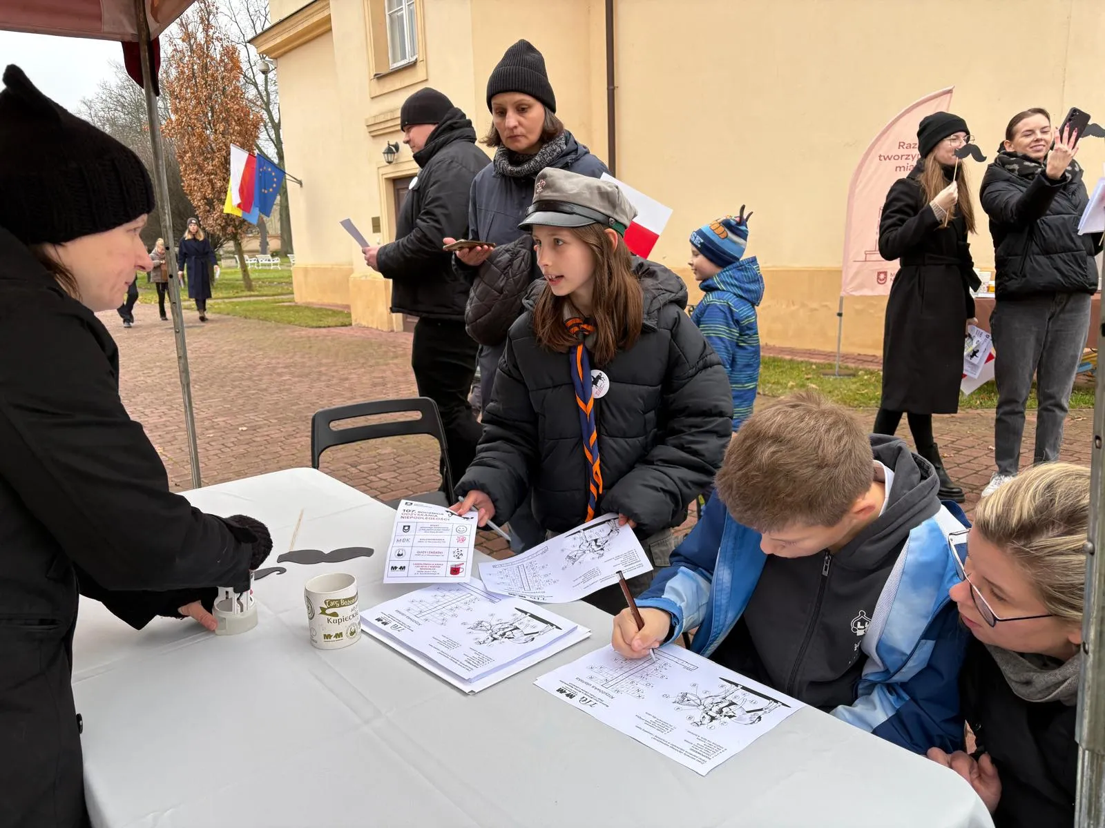 Grupa osób przy stoliku na zewnątrz w chłodny dzień. Dzieci i dorośli w kurtkach i czapkach wypełniają kartki papieru. W tle budynek, flagi i osoby robiące zdjęcia.