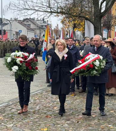 Trzy osoby niosące wieńce kwiatowe w biało-czerwonych barwach idą po kamiennej nawierzchni. Za nimi stoi grupa ludzi z flagami i żołnierze w mundurach. W tle jesienne drzewa i budynki. Dzień, na zewnątrz.