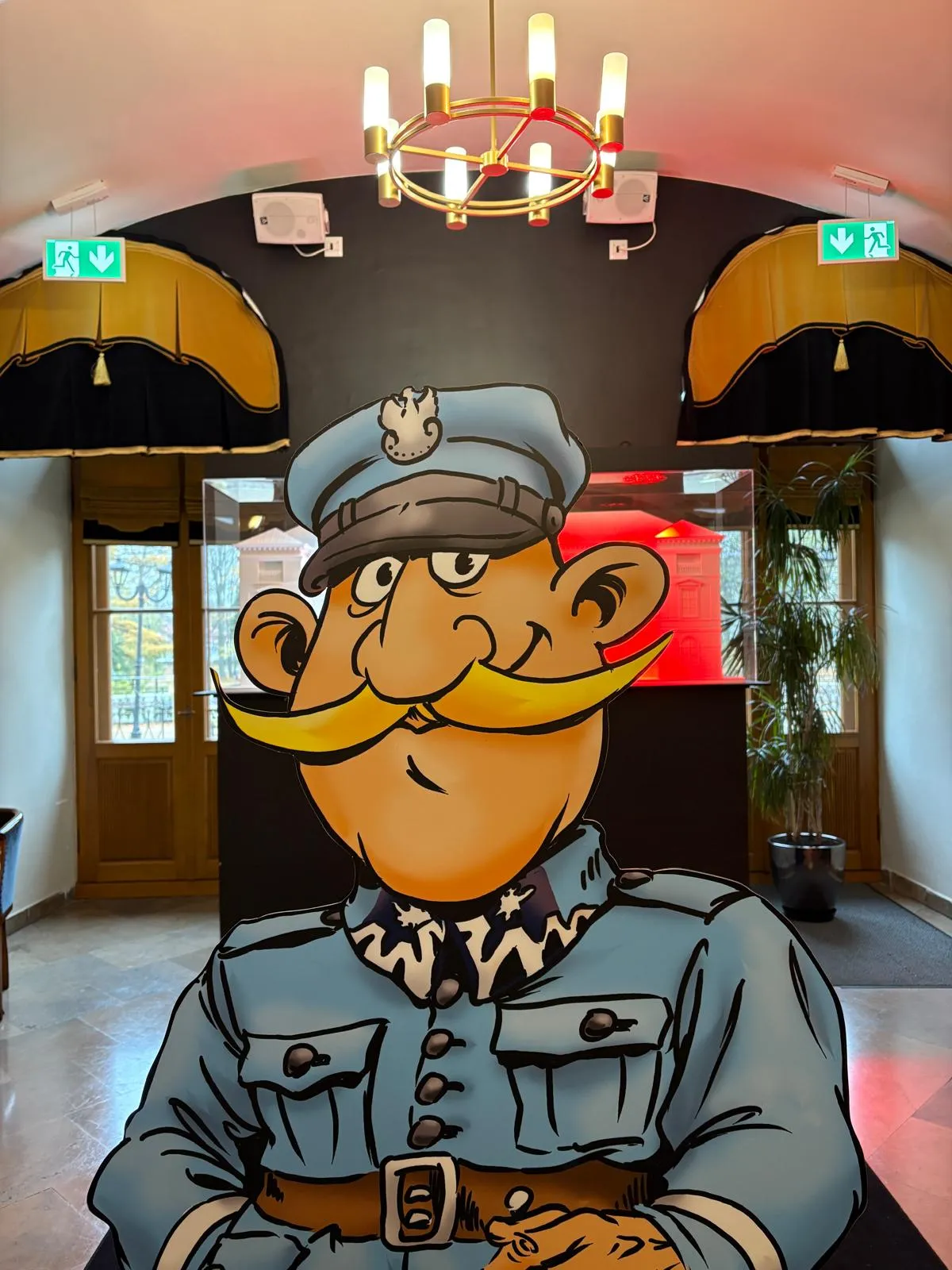 Kartonowa postać żołnierza z dużym żółtym wąsem i niebieskim mundurem stoi w jasnym wnętrzu z dwoma drewnianymi drzwiami, zielonymi znakami ewakuacyjnymi na zwartym, ciemnym tle i wiszącym złotym żyrandolem.