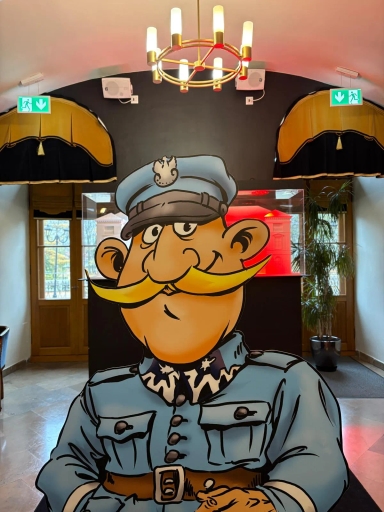 Kartonowa postać żołnierza z dużym żółtym wąsem i niebieskim mundurem stoi w jasnym wnętrzu z dwoma drewnianymi drzwiami, zielonymi znakami ewakuacyjnymi na zwartym, ciemnym tle i wiszącym złotym żyrandolem.