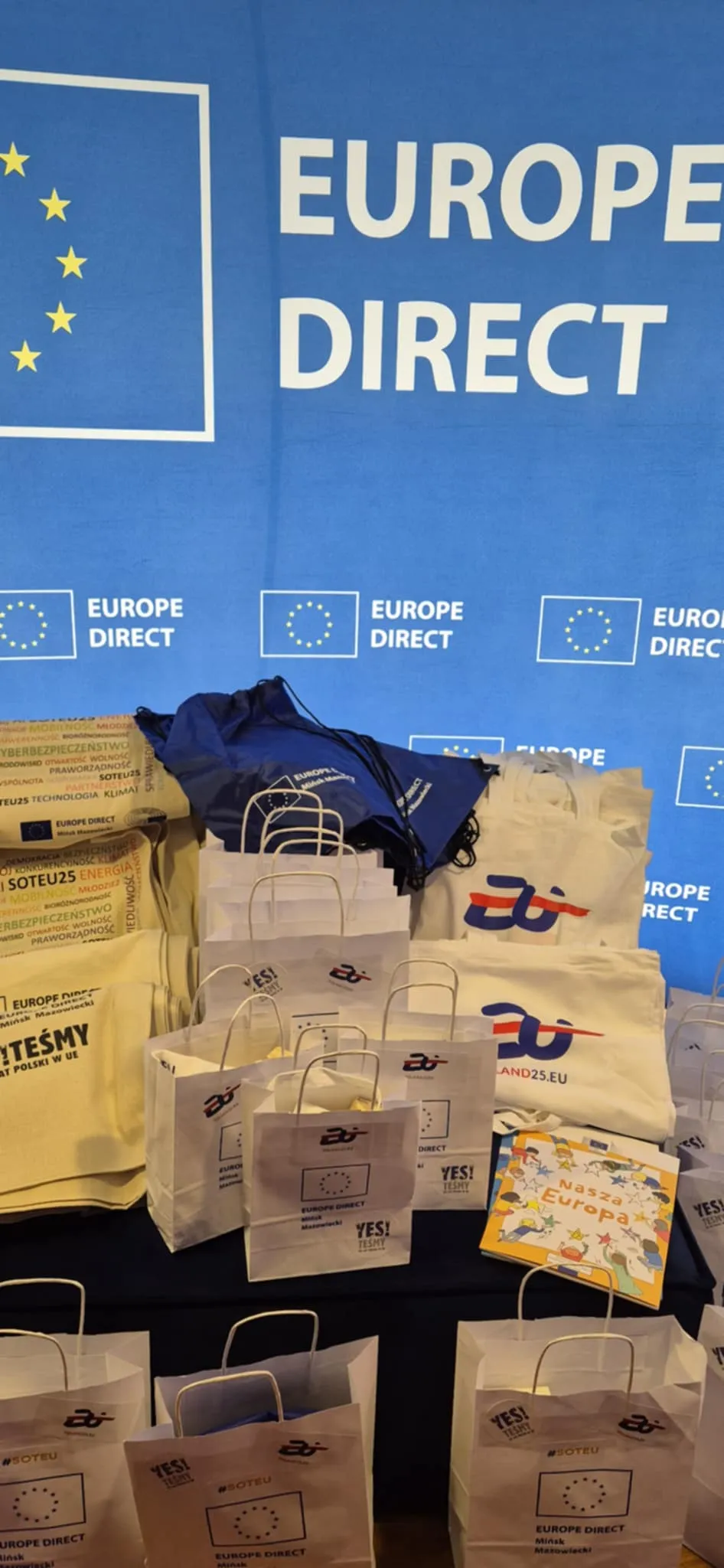 Niebieskie tło z białym napisem EUROPE DIRECT i ikoną gwiazd Unii Europejskiej, na pierwszym planie białe i beżowe torby oraz plecaki z logo Europe Direct, ustawione na stole.
