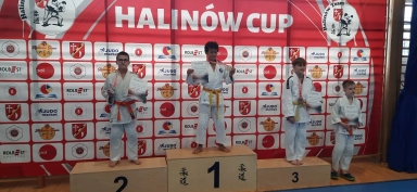 Czterech chłopców w białych judogach stoi na podium z nagrodami i dyplomami na tle czerwono-białej ścianki z logo i napisem HALINÓW CUP.