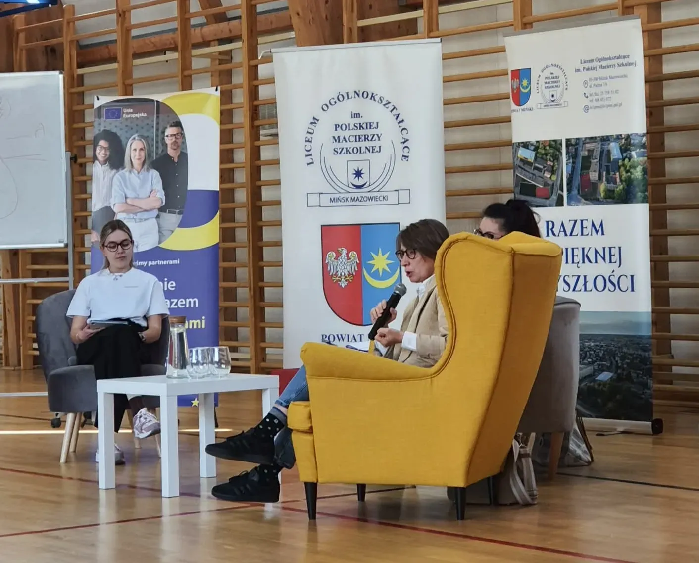 Trzy osoby siedzą na krzesłach w sali gimnastycznej przed drewnianymi drabinkami. Kobieta w żółtym fotelu trzyma mikrofon i mówi. Druga osoba siedzi obok w szarym fotelu. Trzecia osoba czyta notatki przy białym stoliku z dzbankiem i szklankami. Za nimi stoją pionowe banery.