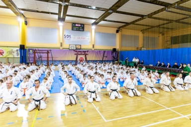 W dużej sali sportowej kilkadziesiąt osób w białych kimonach siedzi na kolanach na drewnianej podłodze i niebieskich matach. Na pierwszym planie kilka osób z czarnymi pasami. W tle sędziowie i widzowie siedzą za stołem z nagrodami. Wnętrze oświetlone, ściany żółte i niebieskie.