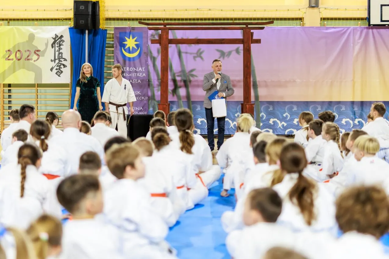 Grupa dzieci i młodzieży w białych kimono siedzi na niebieskich matach w sali gimnastycznej, z przodu stoi mężczyzna mówiący do mikrofonu, kobieta i mężczyzna w kimono obok drewnianej bramy torii, za nimi banery z japońskimi symbolami i napisami.