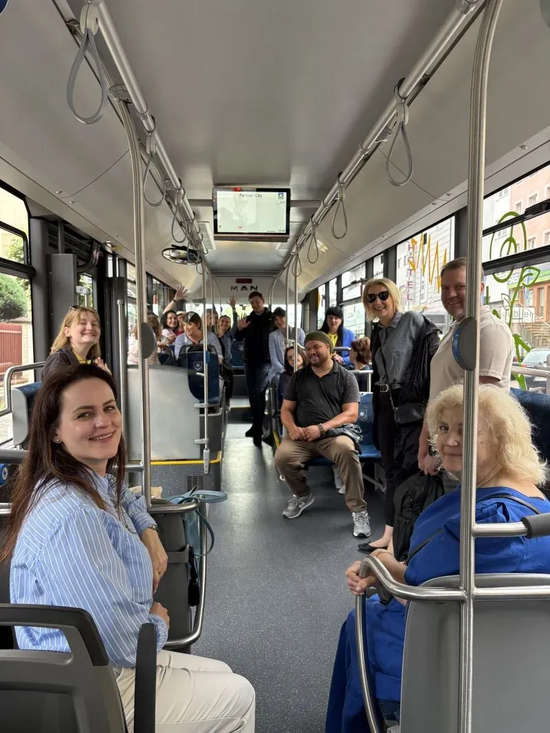 Grupa pasażerów siedzi i stoi wewnątrz autobusu miejskiego.