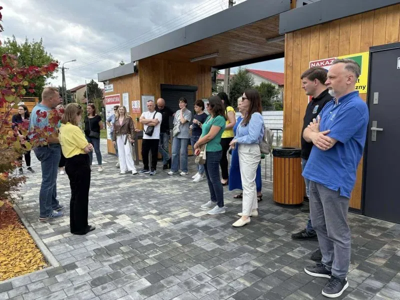 Grupa osób stoi na chodniku przed drewnianą budką lub pawilonem.